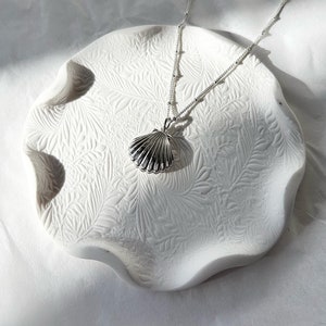 May include: A silver seashell pendant necklace rests on a white, textured, ceramic dish. The dish has a scalloped edge and a leaf pattern. The necklace chain is delicate with small beaded accents.