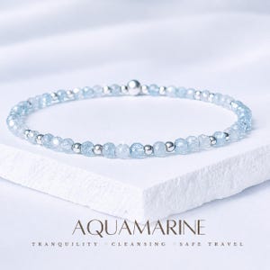 May include: A fine aquamarine bracelet with small, faceted, pale blue beads and silver-coloured metal beads. The bracelet is presented on a white surface, with the word "AQUAMARINE" and other text below.