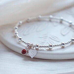 May include: A silver beaded bracelet with a small red gemstone and a heart charm engraved with the letter "S".