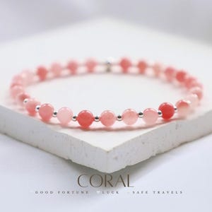 Könnte beinhalten: Ein rosafarbenes Korallenarmband mit silbernen Zwischenstücken. Das Armband liegt auf einer weißen Oberfläche mit dem Text "CORAL GOOD FORTUNE LUCK SAFE TRAVELS".