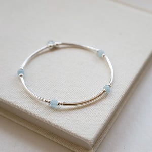 May include: A delicate silver bracelet with four light blue aquamarine beads. The bracelet is curved and sits on a light beige surface. The beads are evenly spaced around the bracelet.