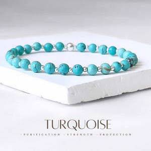 May include: Turquoise beaded bracelet with silver accents. The bracelet is on a white surface. The text "TURQUOISE" is below the bracelet. The text "PURIFICATION STRENGTH PROTECTION" is below the word "TURQUOISE".