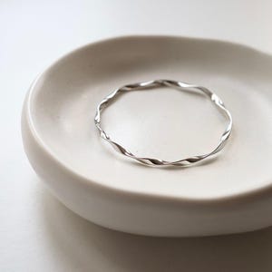 May include: A silver-coloured, twisted metal bangle bracelet. The bracelet has a wavy, textured design and is displayed on a white, shallow dish. The bangle is a simple, elegant piece of jewellery.