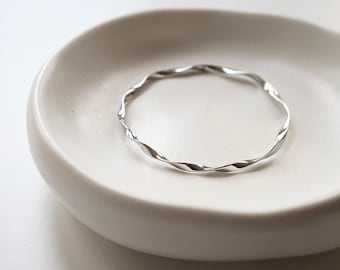 May include: A silver-coloured, twisted metal bangle bracelet. The bracelet has a wavy, textured design and is displayed on a white, shallow dish. The bangle is a simple, elegant piece of jewellery.