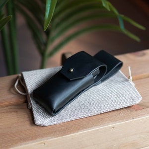 May include: A black leather case with a flap closure and a brass button. The case is sitting on a light brown linen pouch.