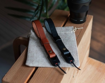 Op de afbeelding: Twee camerabandjes, een van bruin leer en een van zwart leer, liggen op een lichtbruin oppervlak. Beide bandjes hebben een metalen sluiting aan het einde.