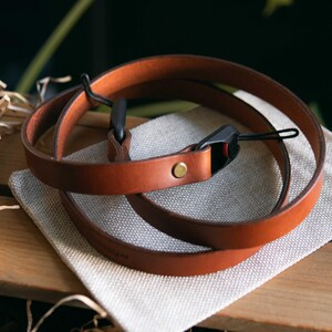 May include: A tan leather camera strap featuring a smooth finish, a single brass-colored metal rivet, and black plastic attachment hardware with thin cord loops for securing to a camera body. The strap is coiled into a circular shape.