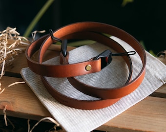 Op de afbeelding: Een beige lederen camerariem met een gladde afwerking, een enkele messingkleurige metalen klinknagel en zwarte kunststof bevestigingsonderdelen met dunne koordlussen voor bevestiging aan een camerabehuizing. De riem is in een cirkel opgerold.