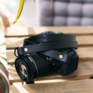 May include: A black leather camera strap attached to a black Canon digital camera. The strap features silver-toned metal rivets securing the leather loops to the camera body. The camera lens is visible, showing the Canon brand name in white text.