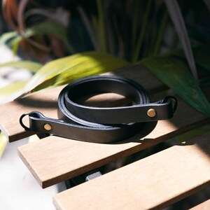 May include: A coiled black leather strap featuring two brass-colored metal rivets and a circular metal ring at one end. The strap is smooth in texture and maintains a consistent width throughout its length, showcasing a simple and functional design.