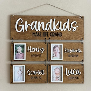 Op de afbeelding: Houten bord met het woord "Grandkids" in witte letters, met de zin "MAKE LIFE GRAND" eronder. Bevat vier kleinere houten platen met foto's en namen: Henry, Elizabeth, Scarlett en Luca, met hun geboortedata.