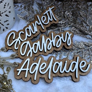 May include: Three wooden Christmas ornaments with names written in white script. The names are "Scarlett", "Gabby", and "Adelaide".