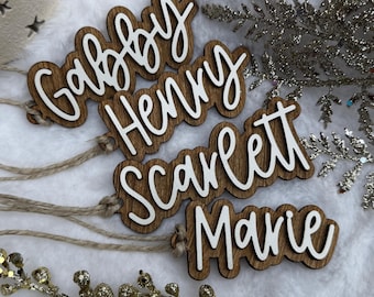Peut inclure: Étiquettes de nom en bois avec des lettres cursives blanches épelant "Gabby", "Henry", "Scarlett" et "Marie". Chaque étiquette est attachée à une ficelle de ficelle naturelle. Les étiquettes sont sur une surface blanche et moelleuse avec des accents décoratifs argentés et dorés.