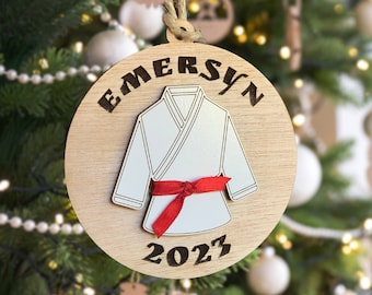 May include: A round wooden Christmas ornament with the name "EMERSYN" and the year "2023" engraved on it. A white karate uniform with a red belt is depicted in the center.