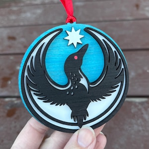 May include: Round wooden ornament featuring a black bird design with a star and red eye on a blue background. The ornament has a white border and a red ribbon for hanging. The bird is a stylized silhouette.