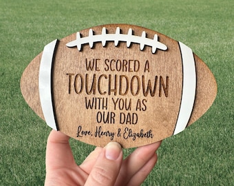 May include: A wooden football-shaped sign with the text "We scored a touchdown with you as our dad. Love, Henry & Elizabeth".