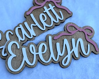 May include: Wooden name signs with the names "Scarlett" and "Evelyn" in white script. Each sign has a pink bow accent and a light brown wood grain background. The signs are layered for a 3D effect.