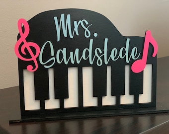 May include: Black wooden piano-shaped sign with a white piano keyboard cutout. The sign has a blue and white cursive inscription that reads "Mrs. Sandstede" and pink musical notes on either side.