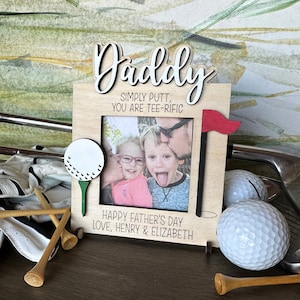 May include: Wooden picture frame with a golf theme. The frame has a golf ball and tee on each side. The text on the frame reads "Daddy Simply Putt, You Are Tee-Rific" and "Happy Father's Day Love, Henry & Elizabeth".