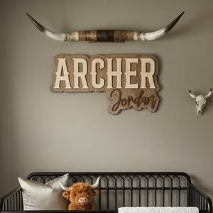 Puede incluir: Un cuarto de ni&ntilde;os con una cuna de metal negro, un peluche marr&oacute;n y un edred&oacute;n blanco. La pared presenta un letrero de madera con el nombre "Archer Jordan", un tapiz de macram&eacute; y una calavera de toro decorativa. Una alfombra de piel de vaca est&aacute; en el suelo.