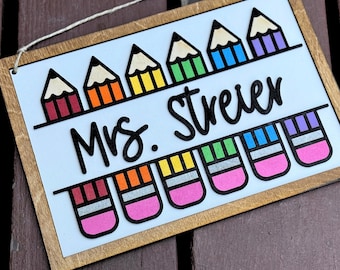 May include: Wooden sign with a white background and a light brown frame. The sign features colorful pencil and eraser graphics. The text "Mrs. Streier" is written in black cursive. A string is attached for hanging.