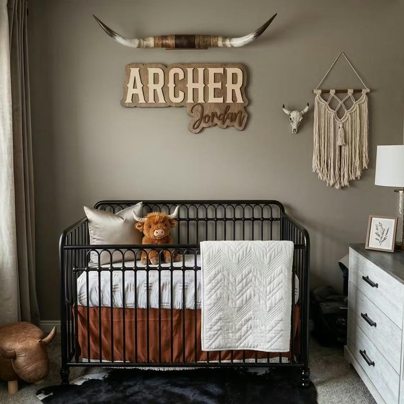 Puede incluir: Un cuarto de ni&ntilde;os con una cuna de metal negro, un peluche marr&oacute;n y un edred&oacute;n blanco. La pared presenta un letrero de madera con el nombre "Archer Jordan", un tapiz de macram&eacute; y una calavera de toro decorativa. Una alfombra de piel de vaca est&aacute; en el suelo.