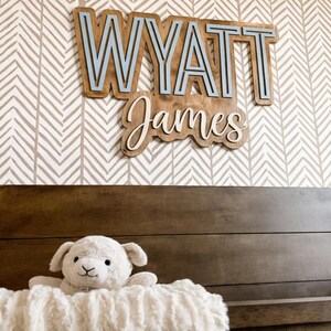 May include: A wooden sign with the name "Wyatt James" in layered lettering, mounted on a wall with a chevron pattern. The name "Wyatt" is in a block font with a light blue interior. A plush lamb sits on a crib.
