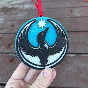 May include: Round wooden ornament with a black bird design, a white star, and a red ribbon for hanging. The bird has a red eye and is set against a light blue background. The ornament is approximately 4 inches in diameter.