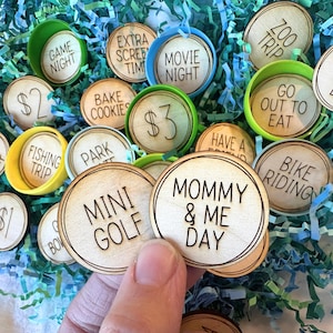 May include: A set of wooden tokens with various activities written on them, such as "Game Night", "Bake Cookies", "Movie Night", "Zoo Trip", "Go Out To Eat", "Fishing Trip", "Park", "Mini Golf", "Mommy & Me Day", "Bike Riding", and "$2" and "$3". The tokens are in a basket filled with green and blue crinkle paper.