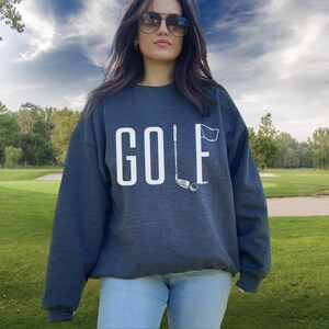 May include: A dark grey sweatshirt featuring the word "GOLF" in white block letters. The "F" is replaced by a golf flag, with a golf club and ball beneath. The sweatshirt is worn by a person wearing sunglasses.