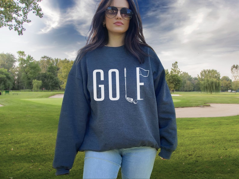 May include: A dark grey sweatshirt featuring the word "GOLF" in white block letters. The "F" is replaced by a golf flag, with a golf club and ball beneath. The sweatshirt is worn by a person wearing sunglasses.