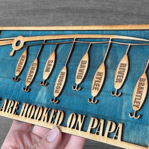 May include: A wooden sign with a blue background and a fishing rod with hooks. The hooks have names on them: Kenzie, Charli, Sam, Hudson, David, Rylee, River, and Brantley. The sign says "We are hooked on Papa".