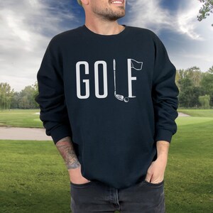 May include: A navy blue sweatshirt featuring the word "GOLF" in white capital letters. The letter "L" is replaced with a golf club and flag graphic. The background shows a golf course with green grass and trees.