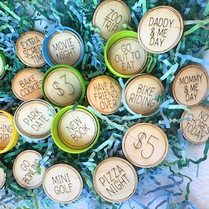 May include: A collection of wooden tokens with various activities and amounts of money written on them. The tokens are in a green and blue shredded paper background. Some of the activities include: "Go Bowling", "Pizza Night", "Bike Riding", "Have a Friend Over", "New Book", "Park Date", "Bake Cookies", "Extra Screen Time", "Movie Night", "Zoo Trip", "Go Out to Eat", "Daddy & Me Day", "Mommy & Me Day", "No Chores Today", "$3", and "$5".