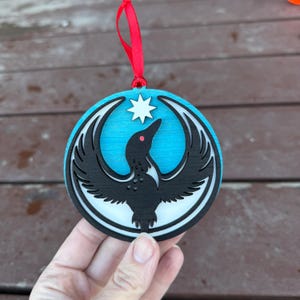 May include: Round wooden ornament featuring a black bird design with a star and red eye on a blue background. The ornament has a white border and is suspended by a red ribbon. The bird has outstretched wings.