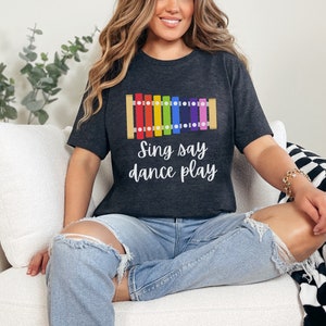 May include: A dark grey t-shirt with a rainbow xylophone graphic and the text "Sing say dance play".