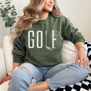 May include: An olive green crew neck sweatshirt featuring the word "GOLF" in white capital letters. A golf club, ball, and flag graphic are next to the word. The sweatshirt is a casual style.