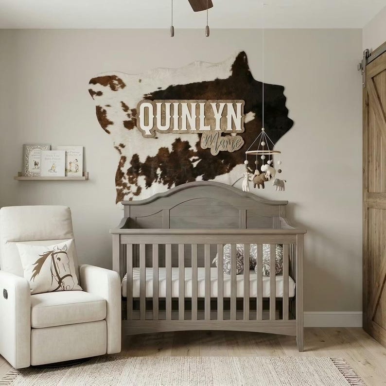 Puede incluir: Un cuarto de beb&eacute; con una cuna gris, un sill&oacute;n reclinable blanco y una decoraci&oacute;n de pared con forma de piel de vaca con el nombre "QUINLYN Marie". Un m&oacute;vil con figuras de animales cuelga sobre la cuna. La habitaci&oacute;n tiene una paleta de colores neutros.