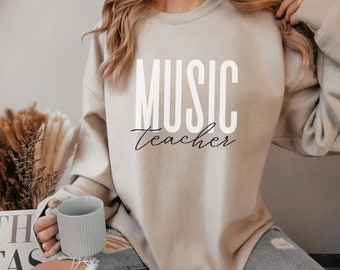May include: Beige sweatshirt with the words "MUSIC teacher" printed in white and black. The sweatshirt is a crew neck style. A person is holding a light blue mug. The person is wearing ripped jeans.