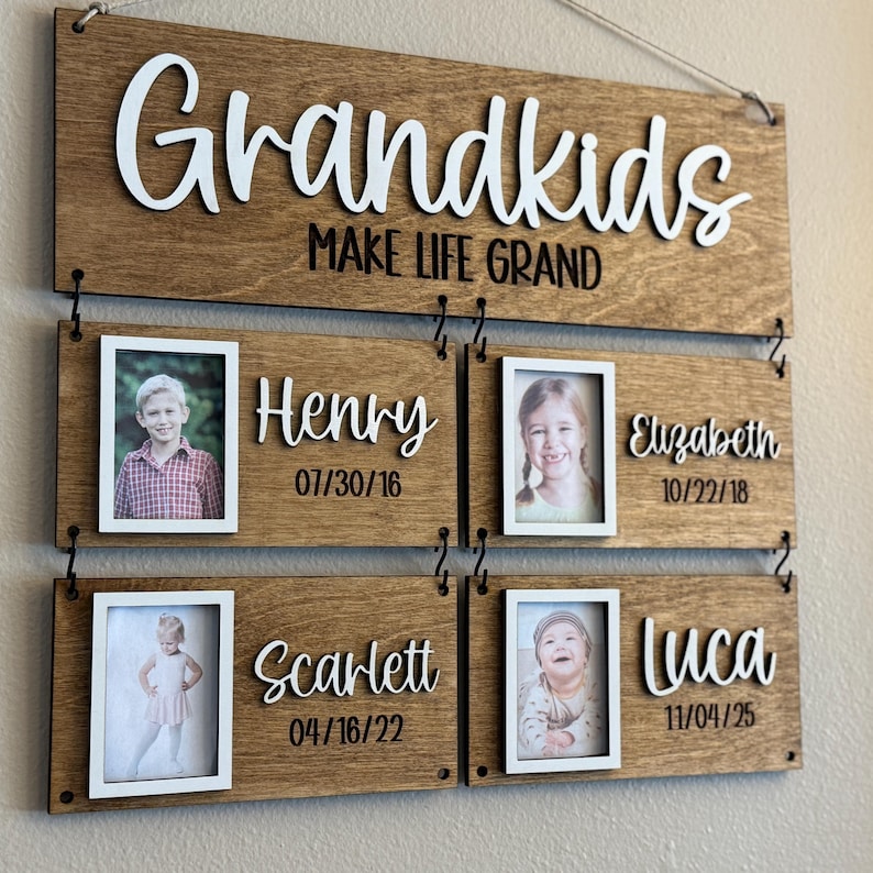 Op de afbeelding: Houten familiebord met het woord "Grandkids" in witte letters. Het bord bevat ruimtes voor foto's en namen: Henry, Elizabeth, Scarlett en Luca, met hun geboortedata. De zin "MAKE LIFE GRAND" is ook opgenomen.