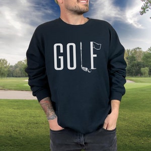 May include: A navy blue sweatshirt featuring the word "GOLF" in white capital letters. The letter "L" is replaced with a golf club and flag graphic. The background shows a golf course with green grass and trees.