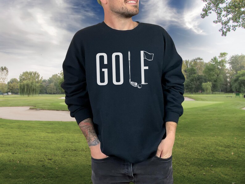 May include: A navy blue sweatshirt featuring the word "GOLF" in white capital letters. The letter "L" is replaced with a golf club and flag graphic. The background shows a golf course with green grass and trees.