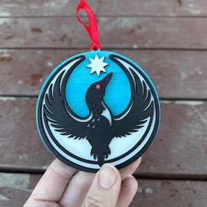 May include: A round, layered wooden ornament with a bird design. The bird is black with a red eye, set against a light blue background. A white star is above the bird. A red ribbon is attached for hanging.