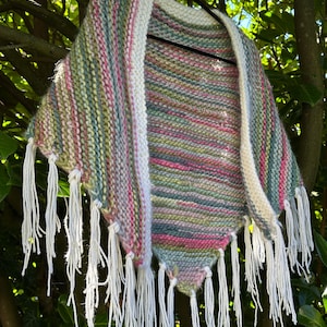 May include: A triangular knit shawl featuring a variegated yarn pattern in shades of pink, green, blue, and yellow. The garment has a thick white knitted border along the top edge and is finished with long white fringe tassels along the bottom edges.