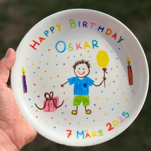 Può includere: Un piatto in ceramica bianca con un'illustrazione disegnata a mano di un bambino che tiene un palloncino giallo, un regalo rosa e due candele. Il testo recita "HAPPY BIRTHDAY OSKAR" in alto e "7 MÄRZ 2025" in basso, circondato da punti colorati.
