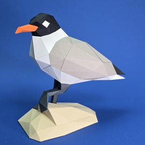 Puede incluir: Escultura geométrica de pájaro de papel con cabeza negra, cara blanca, cuerpo gris y pico naranja. El pájaro está posado sobre una base poligonal beige sobre un fondo azul. La escultura tiene una estética moderna y minimalista.