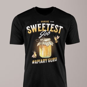 May include: Black t-shirt with a graphic featuring a jar of honey and two bees. The text reads "I HAVE THE SWEETEST Job" above the jar and "#APIARY GURU" below it. The design is in yellow and white.