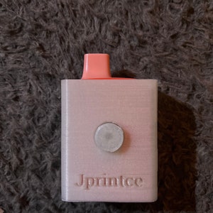 May include: A white 3D printed box with a pink top and a round gray button. The box has the word "Jprintee" printed on the front.