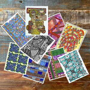 May include: A collection of colorful art prints featuring various abstract patterns and designs. The prints showcase a range of styles, including floral, geometric, and intricate black and white patterns. The artwork is displayed on a rustic wooden surface.