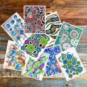 May include: A collection of colorful abstract art prints. Each print features unique patterns of circles, lines, and organic shapes in various colors, including blue, green, pink, and black. The prints are arranged on a wooden surface.